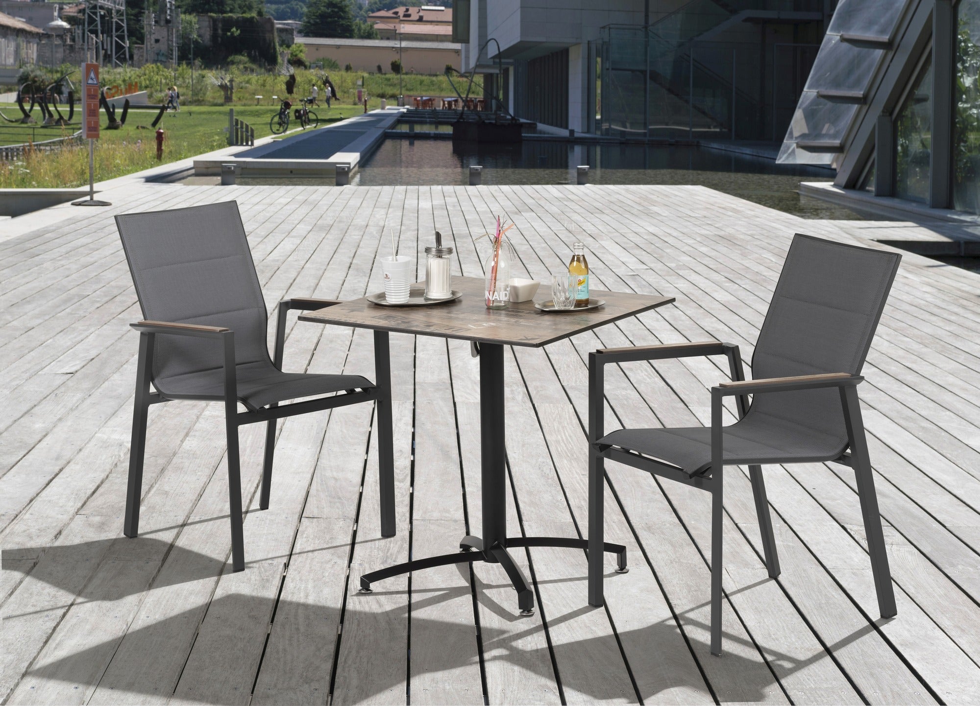 Moderner 'Devin' Gartenstuhl mit strapazierfähigem dunkelgrauem Textilen, leicht gepolstert für zusätzlichen Komfort, mit robustem Metallgestell und Teakholz-Look Polywood-Armstützen, verfügbar bei Gartenmoebelshop.at - Gruppenbild auf einer Terrasse. 2 Armstühle ein Tisch